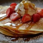 Crêpes légères pliées garnies de fraises fraîches, chantilly et nappées de sirop d'érable, servies sur une assiette sombre avec une touche de sucre glace.