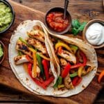 Fajitas au poulet mexicain avec morceaux de poulet grillé, poivrons colorés, tortillas chaudes, accompagnées de guacamole, salsa et crème aigre sur une planche en bois.