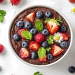 Bowl cake au chocolat garni de fraises, myrtilles, framboises, feuilles de menthe et fleurs comestibles, présenté dans un bol blanc sur une surface claire.