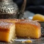 Gâteau de semoule moelleux, décoré de sucre glace, avec une portion coupée, posé sur un plateau en métal avec un thé marocain et des fruits en arrière-plan