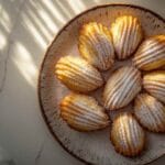Madeleines dorées en forme de coquillage disposées en cercle sur une assiette en céramique, saupoudrées de sucre glace."