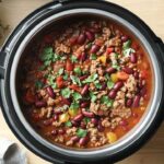 Recette Chili mijoteuse à la viande avec haricots rouges, tomates, poivrons et coriandre fraîche, vu de haut dans une mijoteuse."