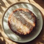 Vue aérienne d’un Gâteau Reine Élizabeth rond nappé de noix de coco râpée, posé sur une assiette blanche, à côté d’un plateau en bois contenant des dattes, le tout baigné d’un doux éclairage naturel.