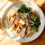 Assiette vue de dessus de pâtes fettuccine Alfredo crémeuses garnies de tranches de poulet grillé, de brocoli croquant et de champignons, sur une planche en bois éclairée par la lumière du jour.