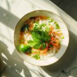 Bol de soupe tonkinoise avec des nouilles de riz, du basilic thaï, des légumes frais, et un quartier de lime, pris en vue de haut dans une cuisine lumineuse.