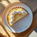 Tarte au citron meringuée avec une tranche manquante, garnie de tranches fines de citron et de petites meringues, présentée sur une assiette blanche posée sur une table en bois clair, éclairage naturel.