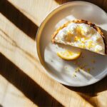 Part de tarte au citron meringuée avec zestes de citron et tranche de citron sur une assiette blanche, vue de dessus avec lumière naturelle et ombres sur une table en bois clair.