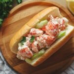 Sandwich guédille au homard avec morceaux de homard en sauce mayonnaise, céleri croquant et feuille de persil, servi sur une planche en bois, accompagné d’un quartier de citron.