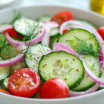 Bol de salade de concombre garni de rondelles de concombre, quartiers de tomates cerises, anneaux d’oignon rouge et brins d’aneth, légèrement assaisonné d’huile d’olive et de poivre.