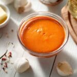 Sauce Buffalo maison orange dans un bol en verre entourée d’ingrédients frais comme du beurre fondu, des piments rouges, de l’ail et des épices sur une table en bois clair.