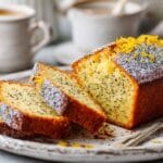 Tranche de cake moelleux au citron avec graines de pavot, décoré de zestes de citron sur une assiette rustique, accompagné de tasses de café en arrière-plan flou.