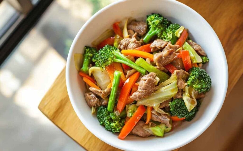 Bol blanc contenant un Chop Suey au porc avec brocoli, carottes, poivrons, et chou, pris en lumière naturelle près d'une fenêtre.