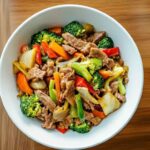 Bol blanc rempli de Chop Suey au porc avec brocoli, poivrons rouges et verts, carottes et oignons, sur une table en bois.