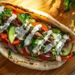 Colis pita garni de viande grillée, concombres, tomates, oignons rouges, persil frais et sauce blanche crémeuse sur un pain pita légèrement grillé, posé sur une planche en bois.