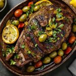 Gigot d’agneau rôti à la marocaine avec olives vertes, tomates cerises, citron grillé et herbes fraîches, présenté dans un plat en terre cuite.