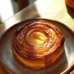 Kouign Amann doré et caramélisé posé sur une assiette en céramique, photographié en lumière naturelle sur une table en bois.