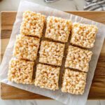 Barres de Rice Krispies maison coupées en carrés, présentées sur une planche en bois recouverte de papier sulfurisé, vue de dessus.