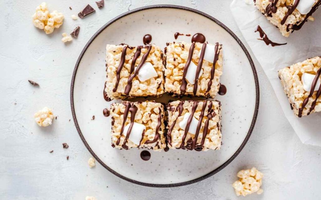 Carrés de Rice Krispies garnis de morceaux de guimauve et nappés de chocolat, servis sur une assiette blanche vue de dessus.