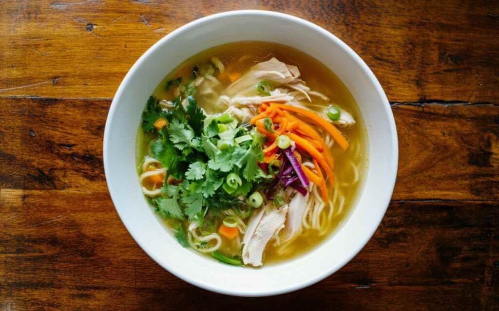 Bol de soupe au poulet avec nouilles, carottes, coriandre fraîche et oignons verts, servi sur une table en bois rustique, vue de dessus.