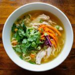 Bol de soupe au poulet avec nouilles, carottes, coriandre fraîche et oignons verts, servi sur une table en bois rustique, vue de dessus.