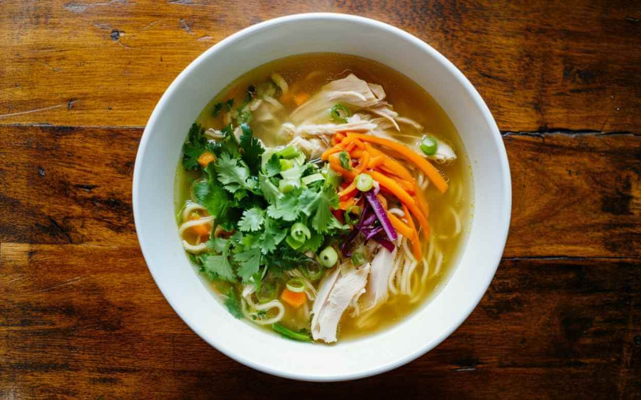 Bol de soupe au poulet avec nouilles, carottes, coriandre fraîche et oignons verts, servi sur une table en bois rustique, vue de dessus.