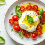 Burrata fraîche nappée d’huile d’olive, entourée de tomates cerises coupées en deux et de feuilles de basilic frais, assaisonnée de poivre noir, présentée dans une assiette blanche sur un fond clair.