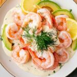 Assiette de crevettes nordiques nappées d’une sauce crémeuse à l’aneth, accompagnées de tranches d’avocat et de citron, présentées sur une assiette blanche avec un fond en bois clair.