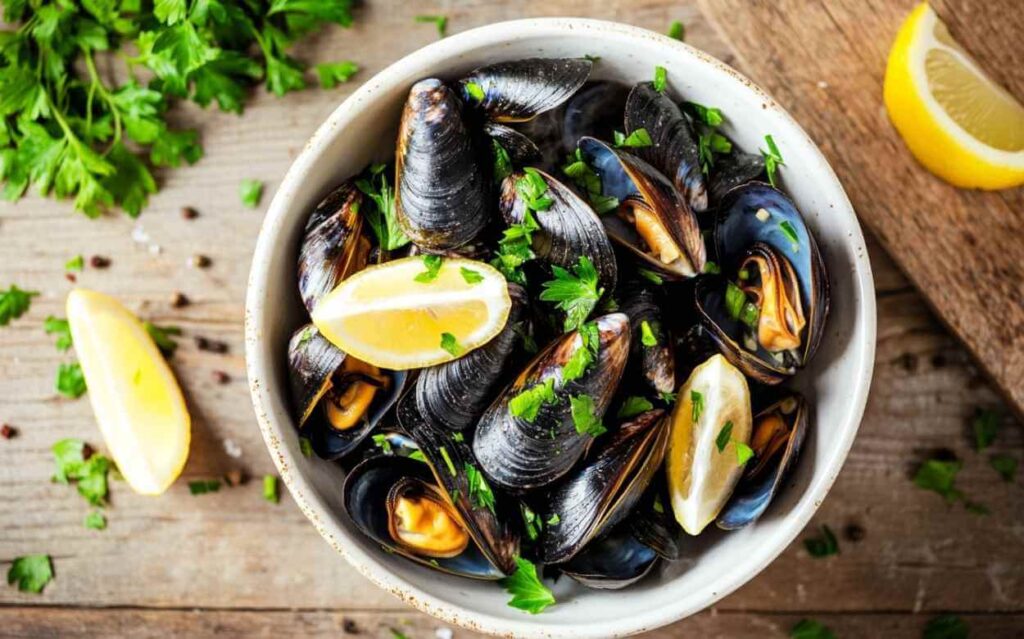 Bol blanc rempli de moules marinières garnies de persil frais et quartiers de citron, posées sur une table en bois rustique avec des herbes et des épices éparpillées autour.