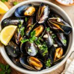 Bol blanc rempli de moules marinières garnies de persil frais et morceaux d’ail, accompagnées de quartiers de citron, posé sur une planche en bois avec une fourchette et une serviette à côté.