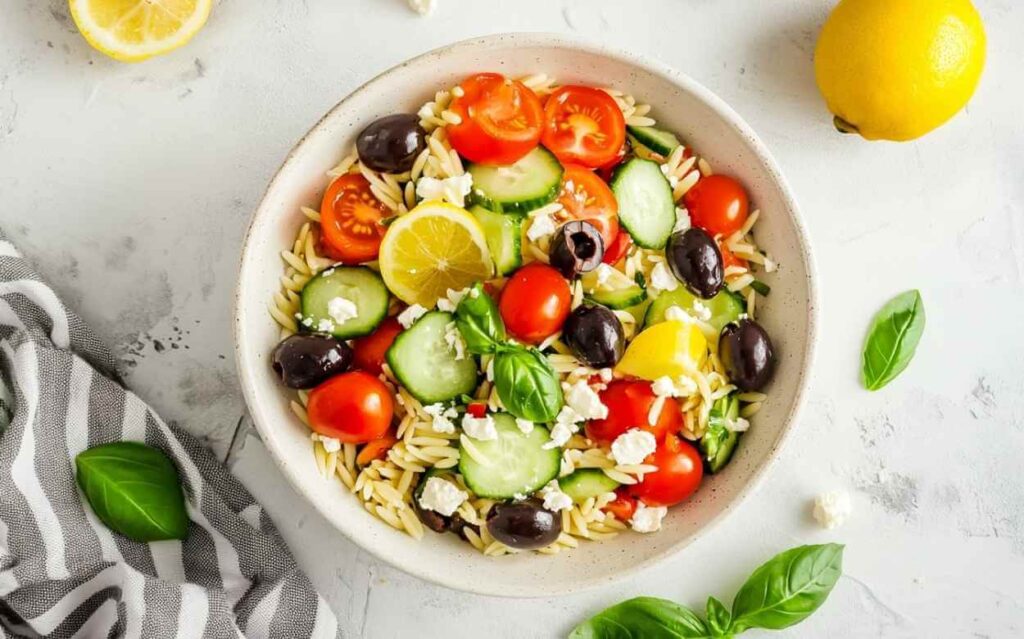 Bol blanc de salade orzo méditerranéenne avec orzo, tomates cerises, tranches de concombre, olives noires, feta émiettée, quartiers de citron et feuilles de basilic frais, sur un plan de travail clair avec linge rayé gris.