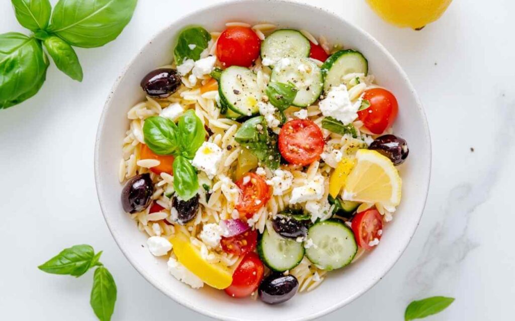 Salade orzo méditerranéenne colorée dans un bol blanc, avec orzo, tomates cerises, concombre, olives noires, feta, feuilles de basilic et quartiers de citron sur fond blanc.