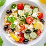 Salade orzo méditerranéenne colorée dans un bol blanc, avec orzo, tomates cerises, concombre, olives noires, feta, feuilles de basilic et quartiers de citron sur fond blanc.