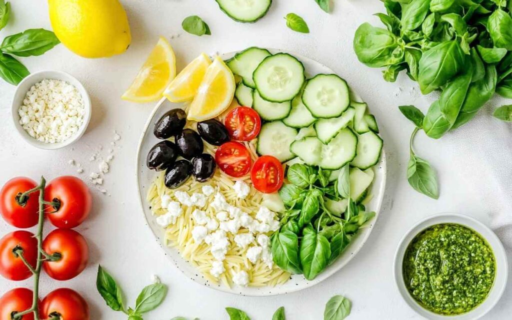 Assiette avec ingrédients séparés pour salade orzo : orzo, feta émiettée, tomates cerises, olives noires, rondelles de concombre, feuilles de basilic, quartiers de citron, accompagnée d’un bol de pesto et de tomates grappes, le tout sur un fond blanc.