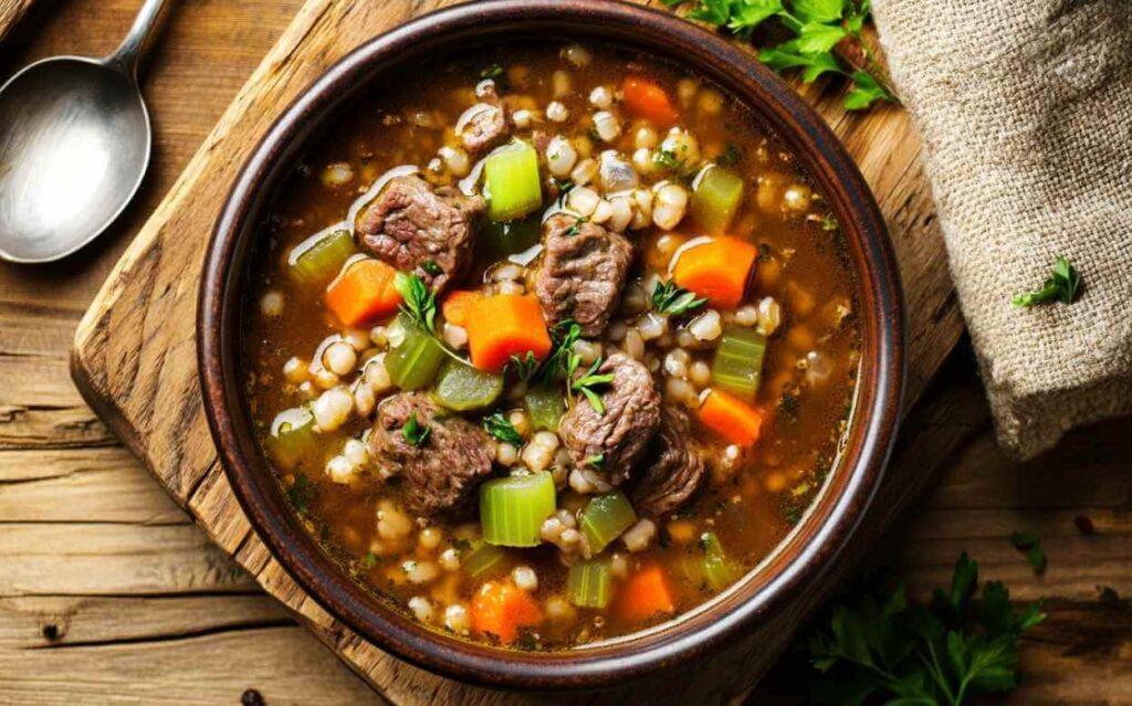 Bol de soupe bœuf et orge avec morceaux de viande, carottes, céleri et herbes fraîches, présenté sur une planche en bois rustique avec une cuillère en métal à côté.