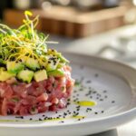 Tartare de thon frais et avocat coupé en dés, joliment dressé en cercle sur une assiette blanche, garni de pousses et graines de sésame noir, avec un filet d'huile d'olive, en arrière-plan cuisine lumineuse.