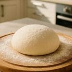 Boule de pâte à pizza maison levée sur une planche en bois, saupoudrée de farine, dans une cuisine moderne lumineuse.