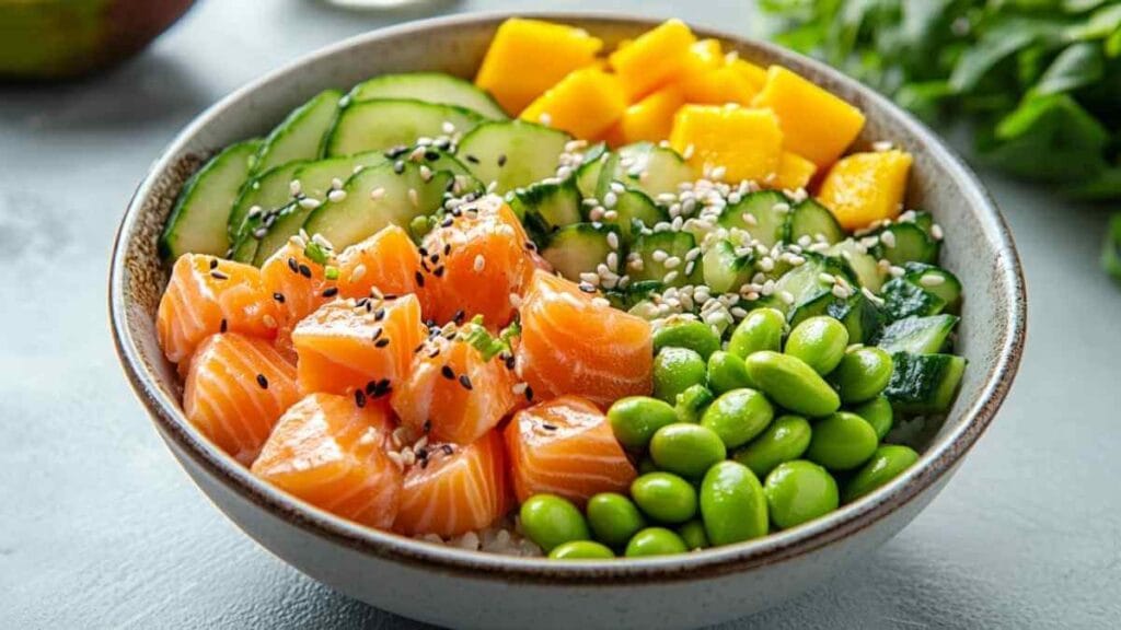 Bol de poke bowl avec cubes de saumon frais, edamame, dés de mangue, concombre en tranches, le tout garni de graines de sésame sur un fond clair.
