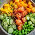 Poke bowl coloré avec cubes de saumon frais, dés de mangue, tranches de concombre, edamame et graines de sésame, présenté dans un bol sur un fond gris.
