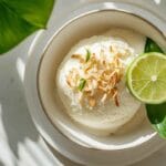 Bol de sorbet noix de coco garni de copeaux de noix de coco grillés et d’une tranche de citron vert, présenté sur une assiette blanche avec une feuille verte, éclairage naturel.