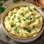 Bol en céramique rempli de purée de pommes de terre maison garnie de persil frais haché, posé sur une nappe en lin beige et une table en bois rustique, avec une cuillère en bois à côté.