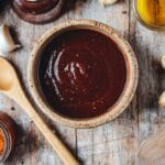 Sauce barbecue maison épaisse dans un bol en céramique, entourée d’épices, gousses d’ail, huile et une cuillère en bois, sur une table en bois rustique.