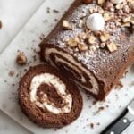 Bûche de Noël au chocolat roulée, garnie de crème, saupoudrée de sucre glace et de noisettes concassées, tranchée sur planche de marbre avec couteau et mini meringues.