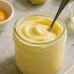 Homemade mayonnaise in a glass jar with a spoon, surrounded by egg yolk, lemon, mustard, vinegar, salt, and pepper on a marble surface.
