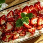 Vue de dessus d’un tiramisu aux fraises dans un plat en verre, décoré de tranches de fraises fraîches, de zestes de citron et de feuilles de menthe, éclairé par une lumière naturelle.