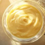 Bol de crème pâtissière lisse et brillante vu de dessus, avec une gousse de vanille et un fouet en arrière-plan dans une cuisine ensoleillée.