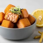 Bouchées de saumon panées et croustillantes servies dans un bol gris, garnies de graines de sésame noir et d'oignon vert, avec un demi-citron.