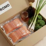 Deux filets de saumon emballés sous vide dans une boîte repas "PROMO" avec du brocoli, des oignons verts et deux petites bouteilles de sauce ou d'huile.