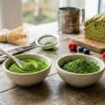 Deux bols de poudre de thé matcha vert vif posés sur une table en bois rustique. Le bol de gauche contient une cuillère en céramique blanche remplie de poudre. En arrière-plan, on aperçoit des outils de pâtisserie, un pain d'épices au matcha, des framboises et des myrtilles.